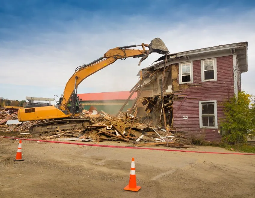 Large yellow excavator demolishing an old two-story residential house in Dallas to clear the lot for new construction